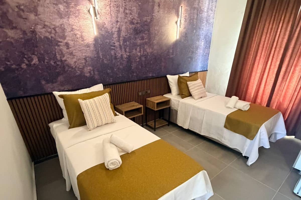 Habitación de hotel con dos camas, una mesita de noche de madera, una lámpara de pared y una ventana con cortinas.