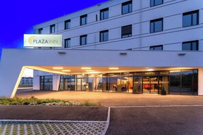 Moderni hotellin sisäänkäynti, jossa on lasijulkisivu, kyltti, josta ilmenee, että kyseessä on Plaza Inn -hotelli, sekä selvästi näkyvä ulkona oleva oleskelualue.