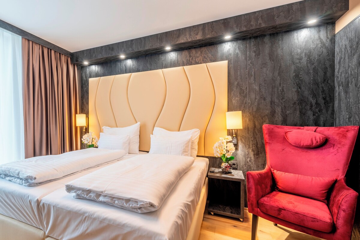 A hotel room with a large bed, two bedside tables, a red armchair, and a view of the outdoors.