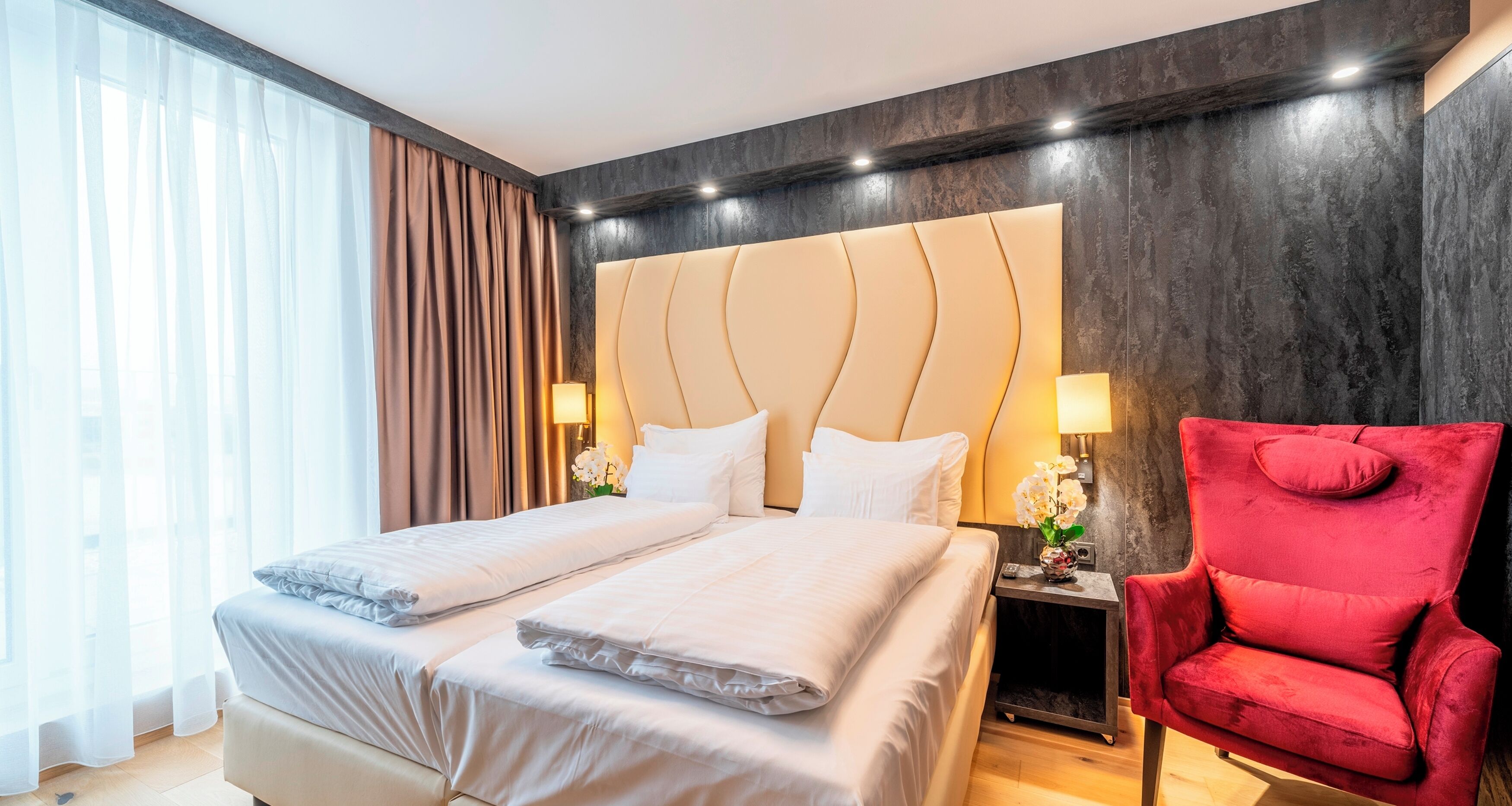 Habitación de hotel con una cama grande, dos mesitas de noche, un sillón rojo y vistas al exterior.