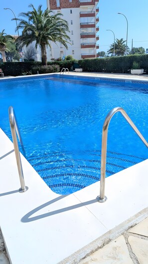Una piscina con barandillas metálicas, rodeada por un deck de baldosas, palmeras y un edificio de varios pisos al fondo.