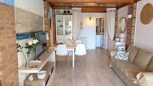 A cozy living room with a sofa, a dining area with a table and chairs, a kitchen visible through an open door, and a brick wall with a painting. - Apartment 'Sea and Charm' with Shared Pool, Wi-Fi, and Air Conditioning (Miami Platja)