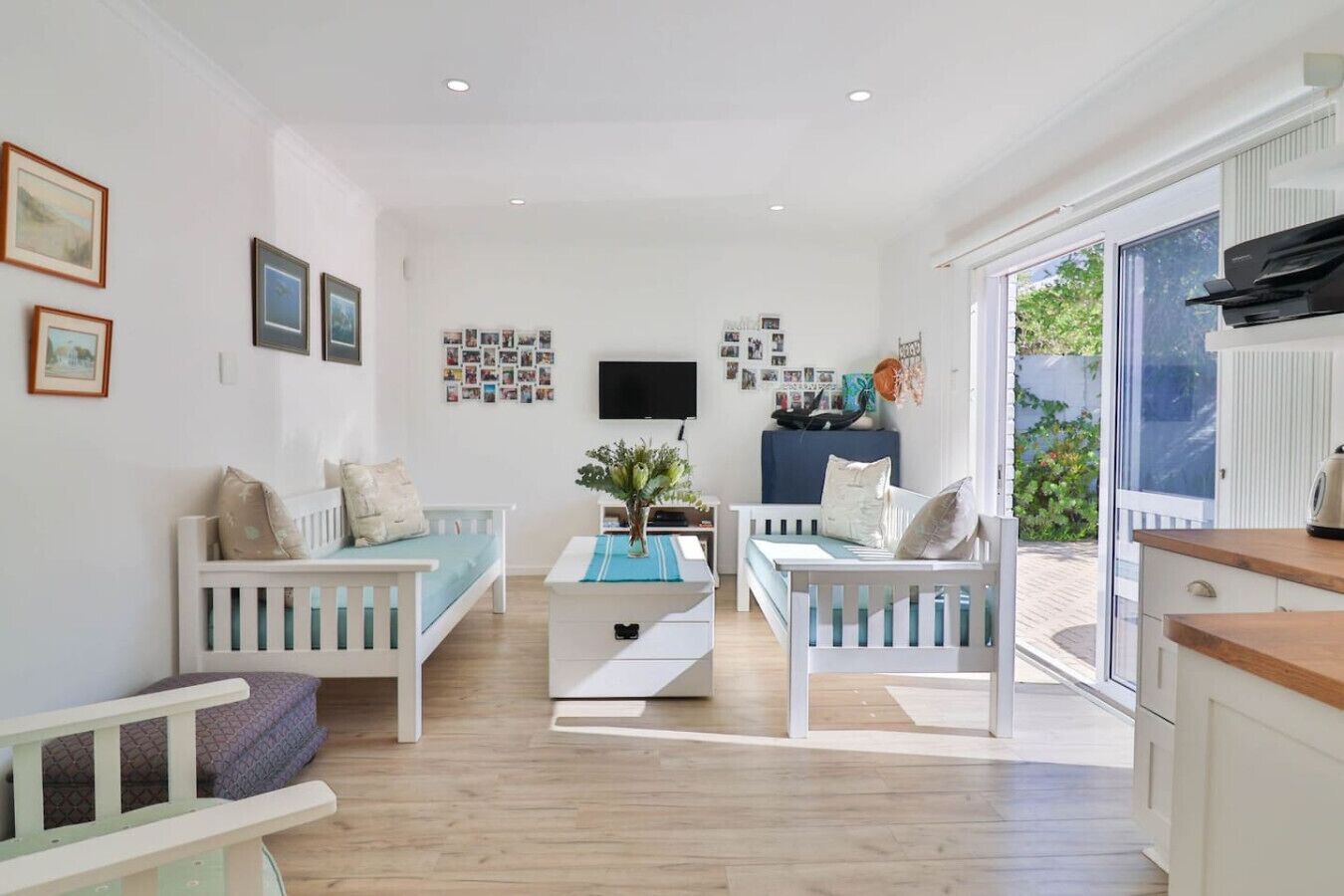Un salon lumineux avec deux canapés blancs, une table basse et un téléviseur. Les murs sont ornés de photos encadrées et une fenêtre offre une vue sur la verdure.