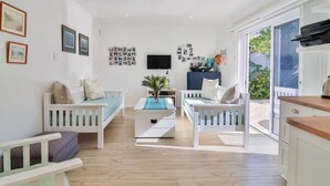 A bright living room with two white sofas, a coffee table, and a TV. There are framed photos on the walls and a window with a view of greenery. - Vonkelwyn | Bright and Breezy by Grotto Beach (Hermanus)
