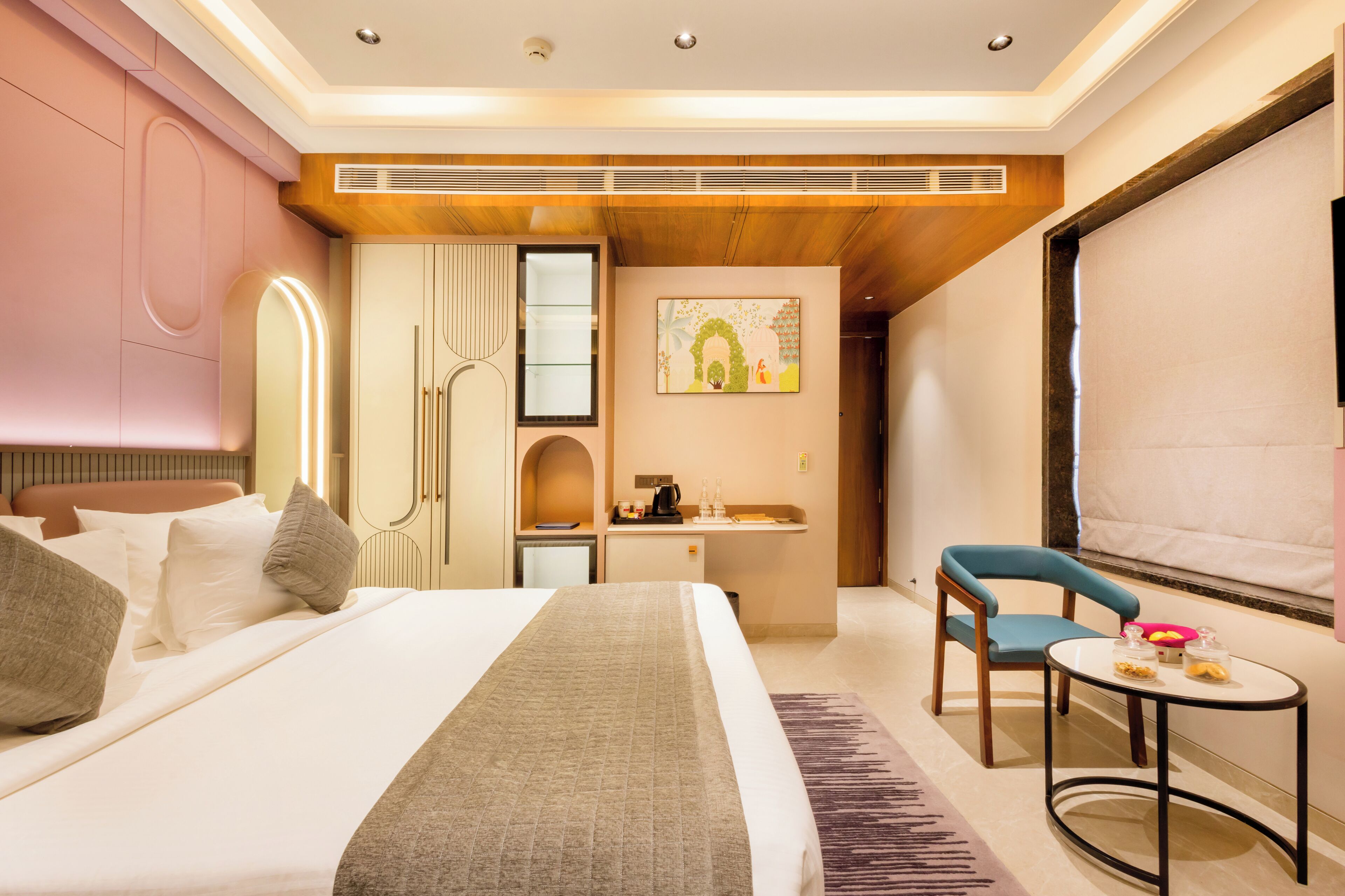 Quarto de hotel com cama, escrivaninha, cadeira, mesa pequena e um quadro na parede.