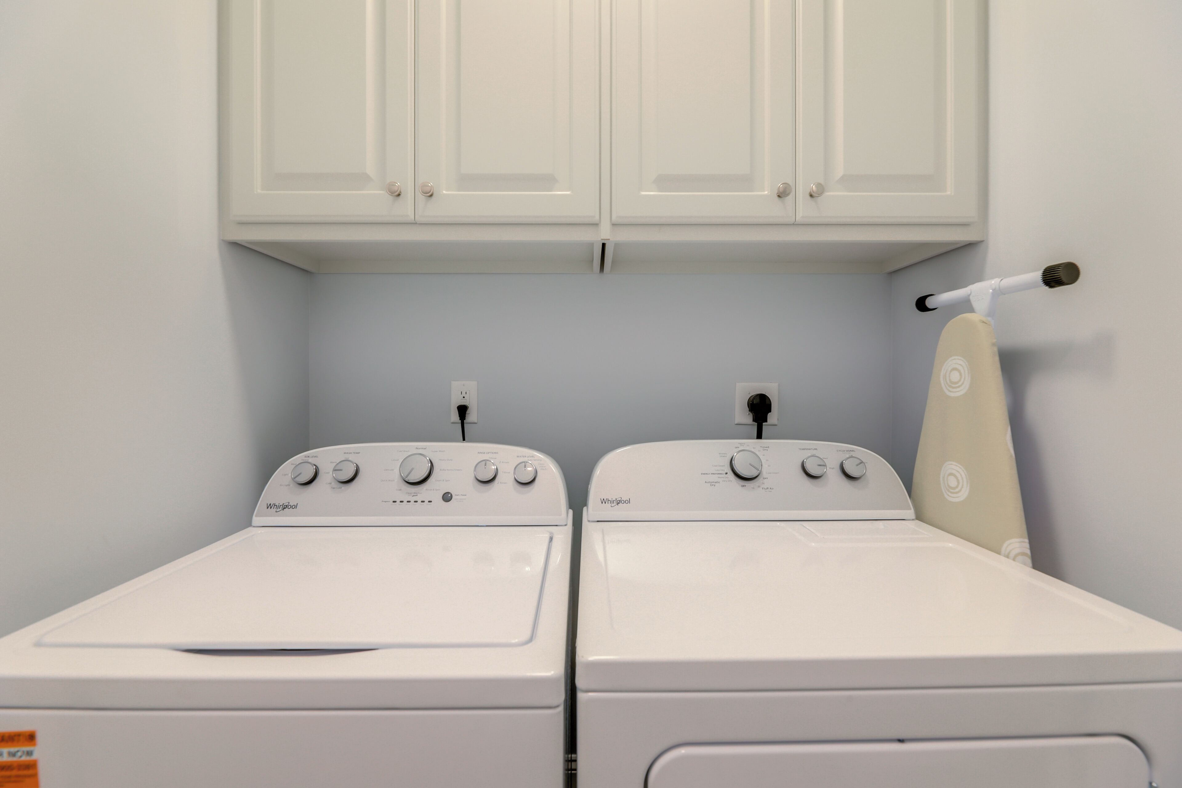 Two white Whirlpool washers with control panels, a white ironing board, and a white cabinet above them.
