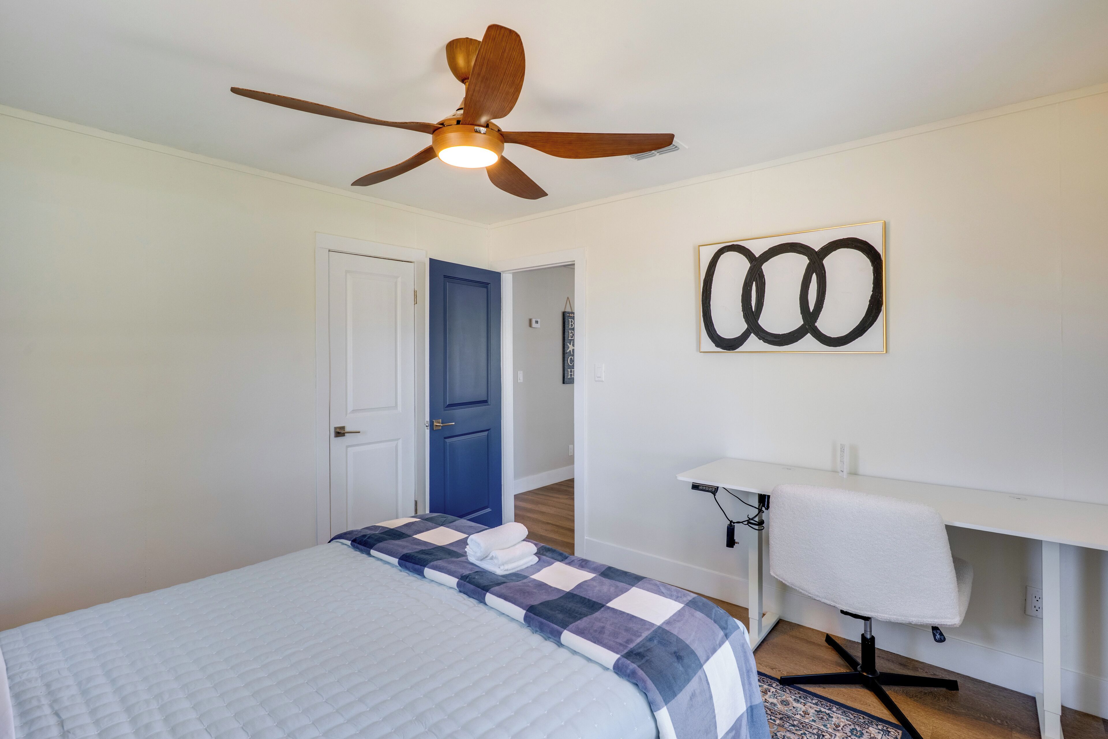 A bedroom with a bed, a ceiling fan, a desk, and a wall art piece.
