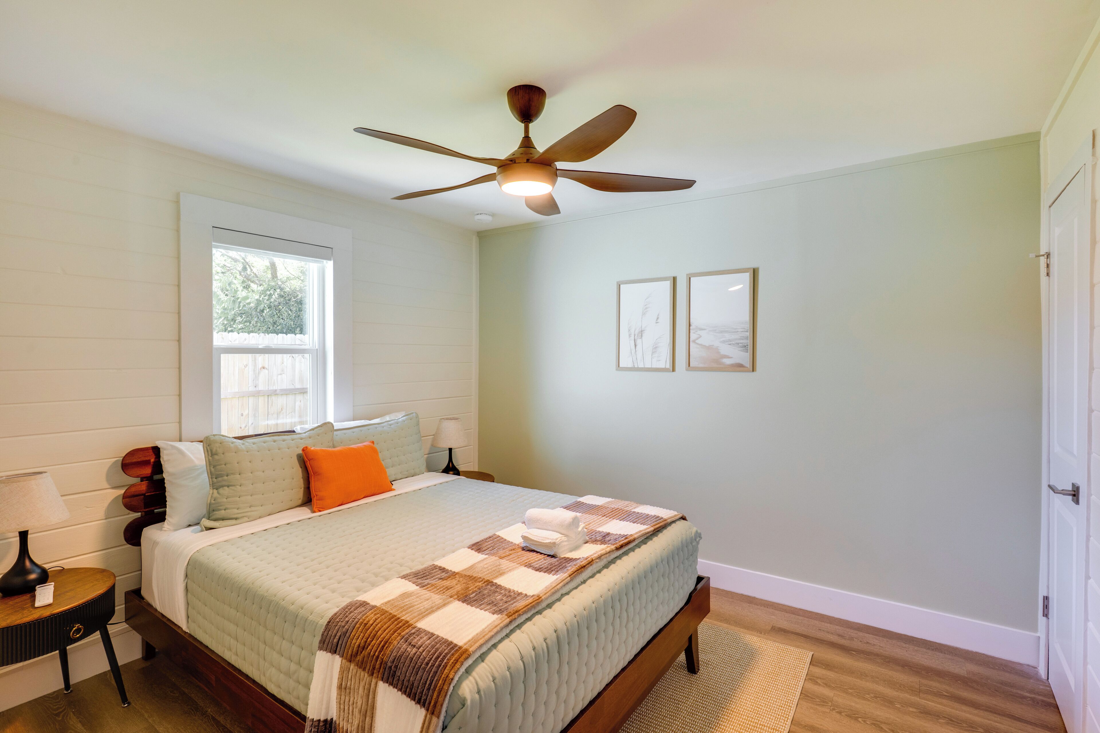 A bedroom with a bed, a ceiling fan, a window, and framed pictures on the wall.