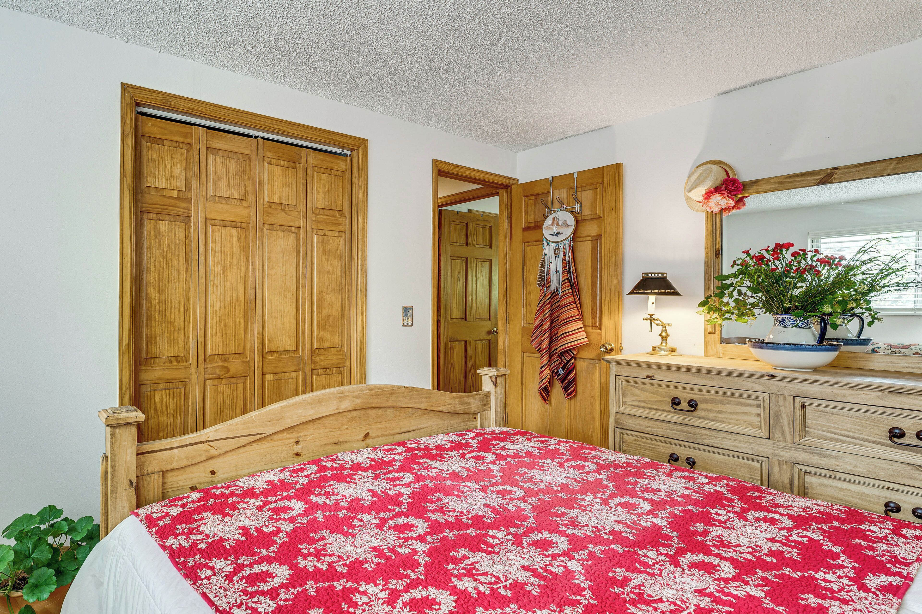 A bedroom with a wooden bed, a red floral bedspread, a wooden dresser, a mirror, and a door leading to another room.