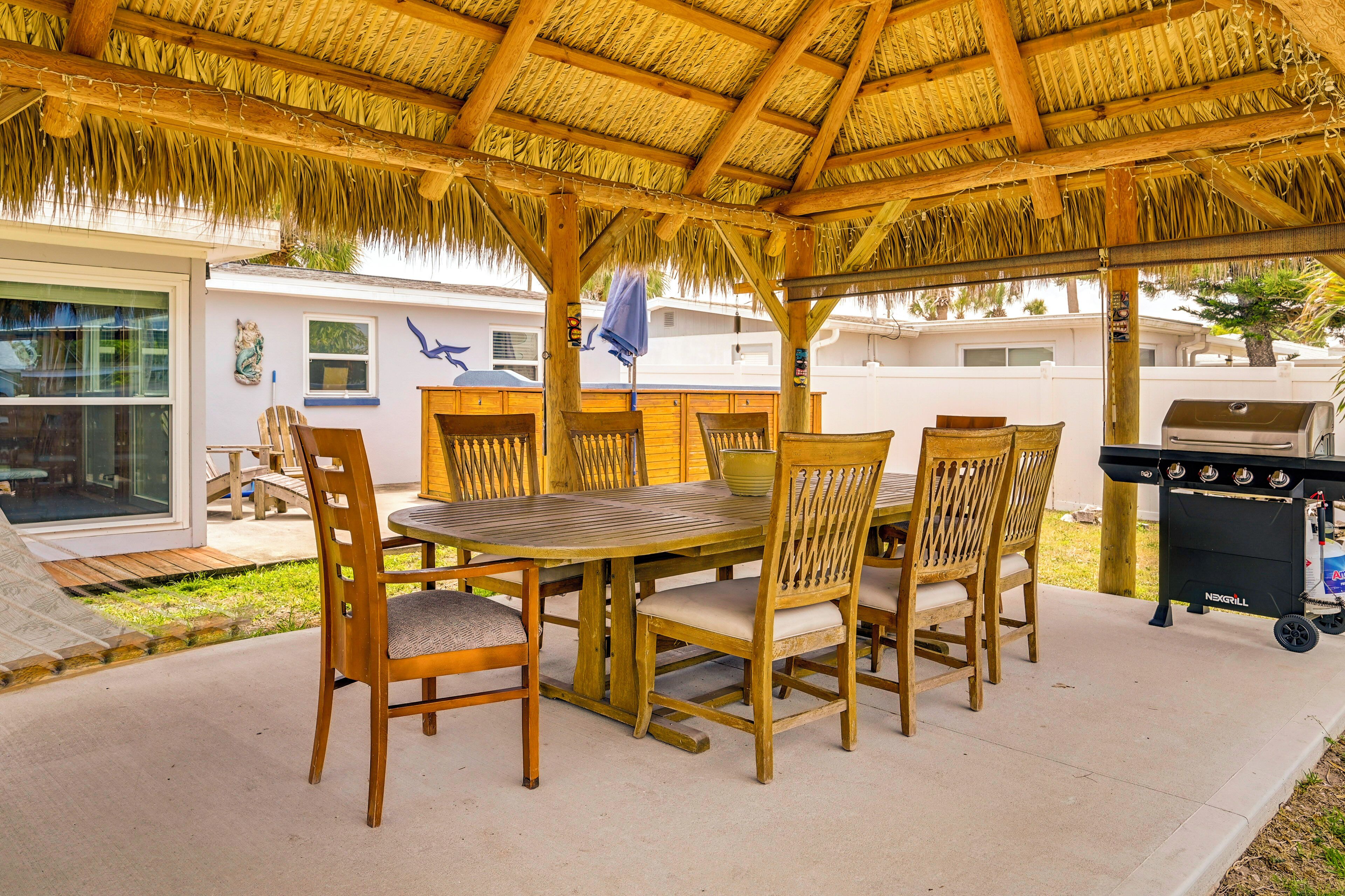 A covered patio with a thatched roof, wooden chairs, a round table, and a grill.