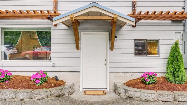 Una casa con puerta blanca, porche de madera y un pequeño jardín con flores rosas.