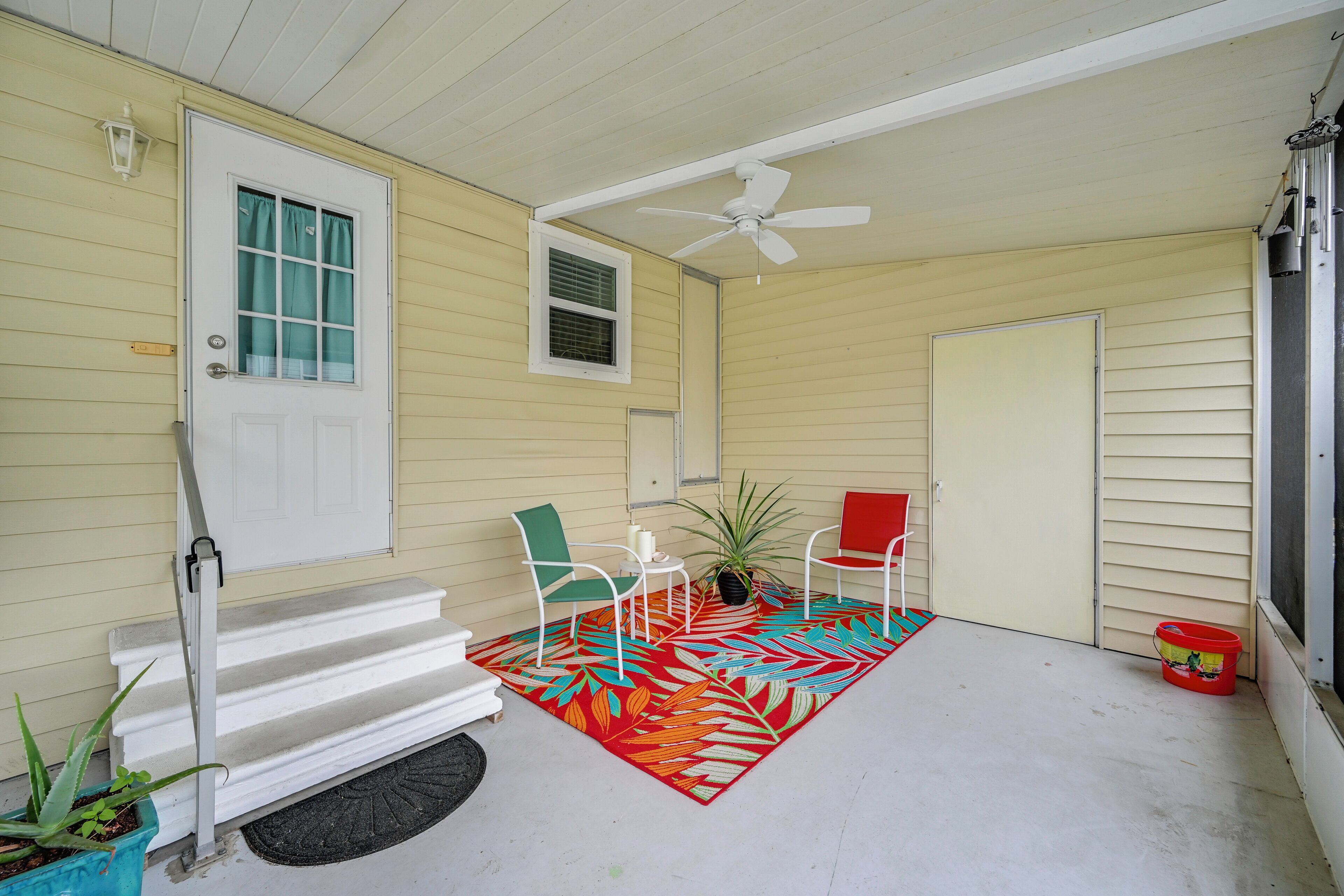 A covered porch with a white door, a ceiling fan, a red and orange patterned rug, two white chairs, a red chair, a potted plant, and a red bucket.