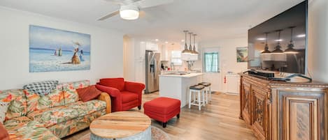 A living room with a sofa, ottoman, and round coffee table, a kitchen with a refrigerator and island, and a dining area with bar stools.