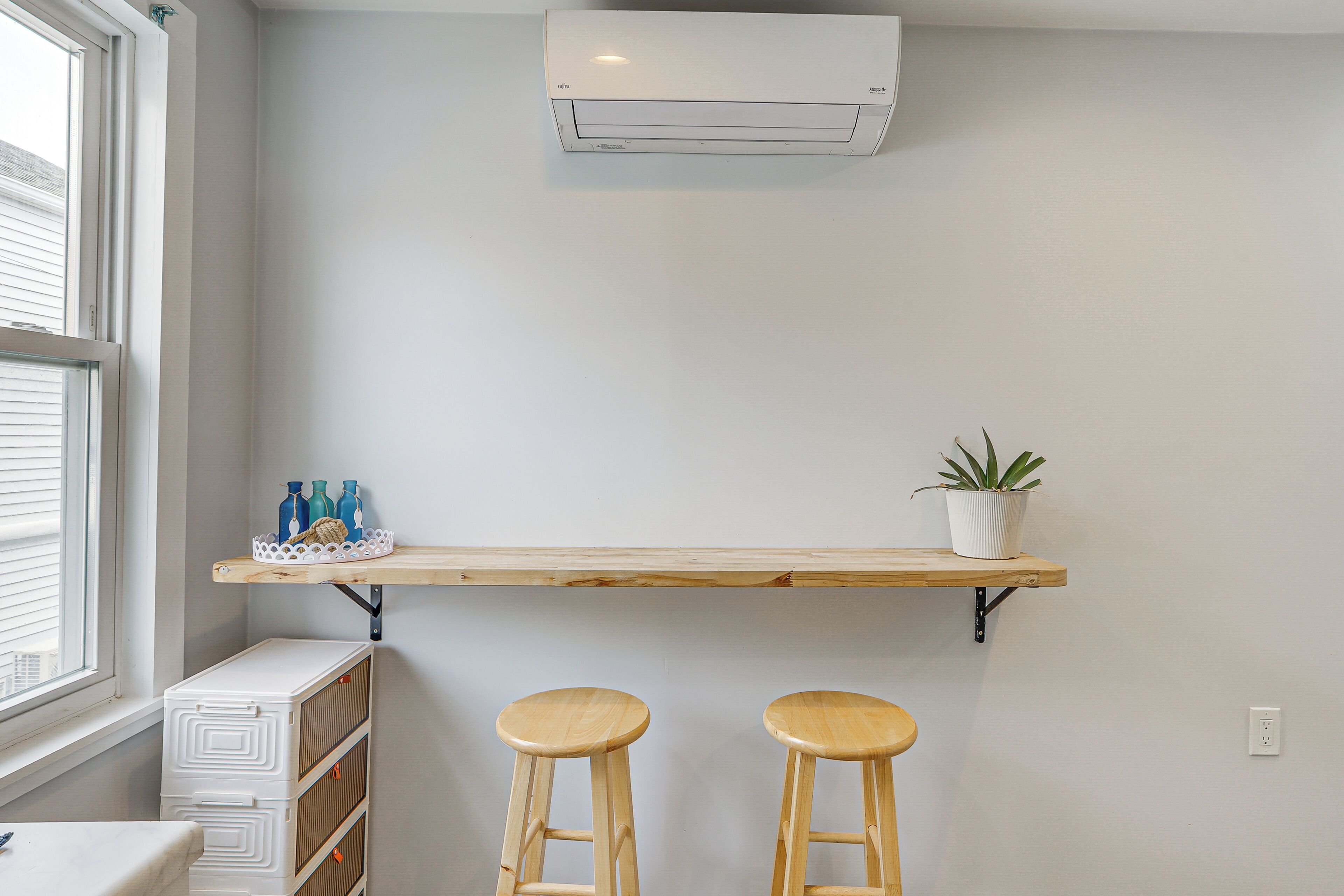 A room with a window, an air conditioner, a shelf with bottles and a plant, and two stools.