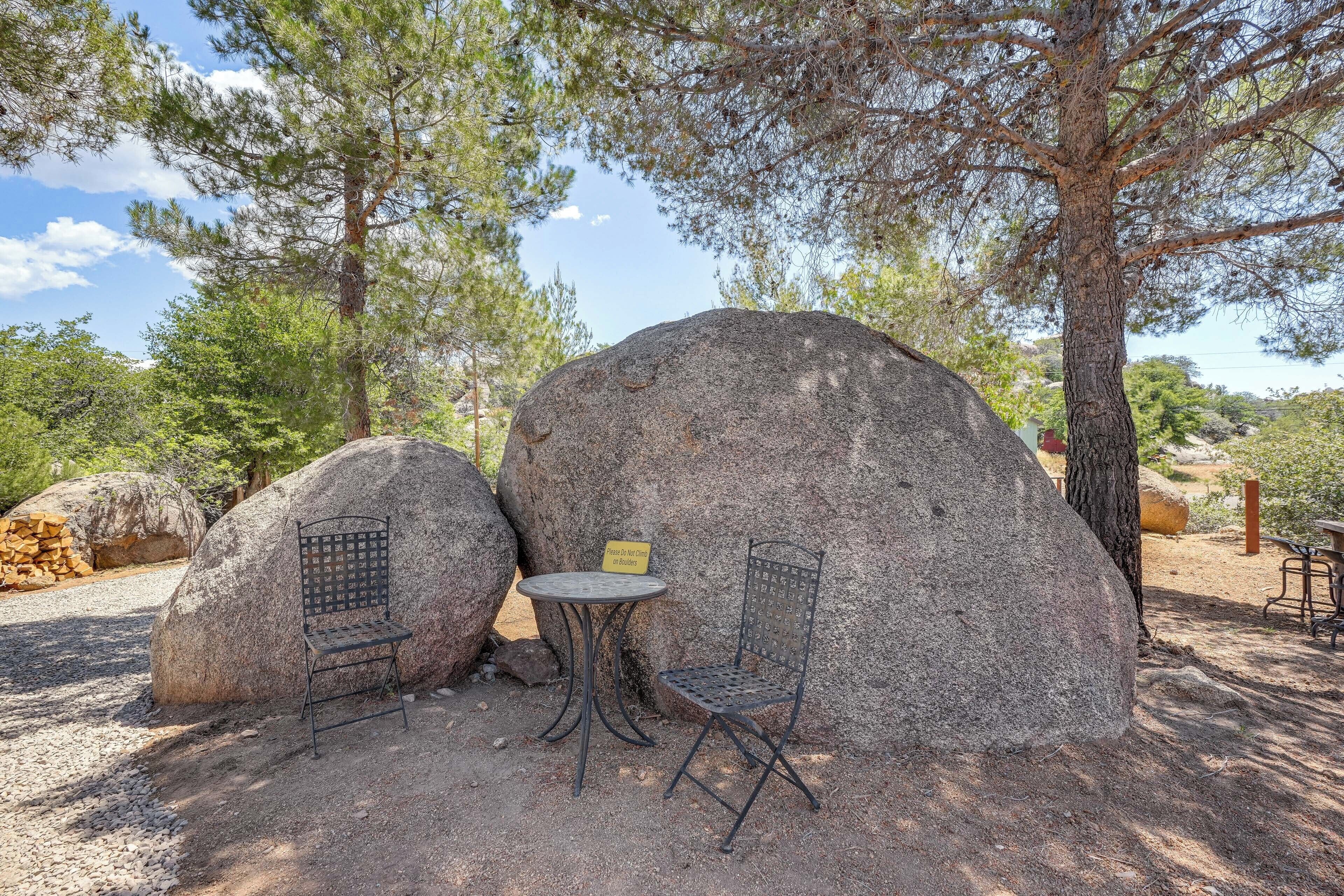 A rustic outdoor seating area with two metal chairs and a small round table, surrounded by large rocks and trees.
