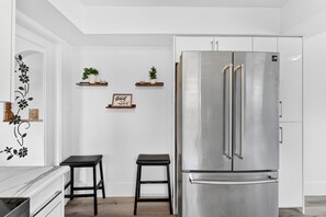 Eine moderne Küche mit einem Edelstahlkühlschrank, zwei schwarzen Hocker, weißen Wänden und dekorativen Regalen mit Pflanzen und einem Schild.