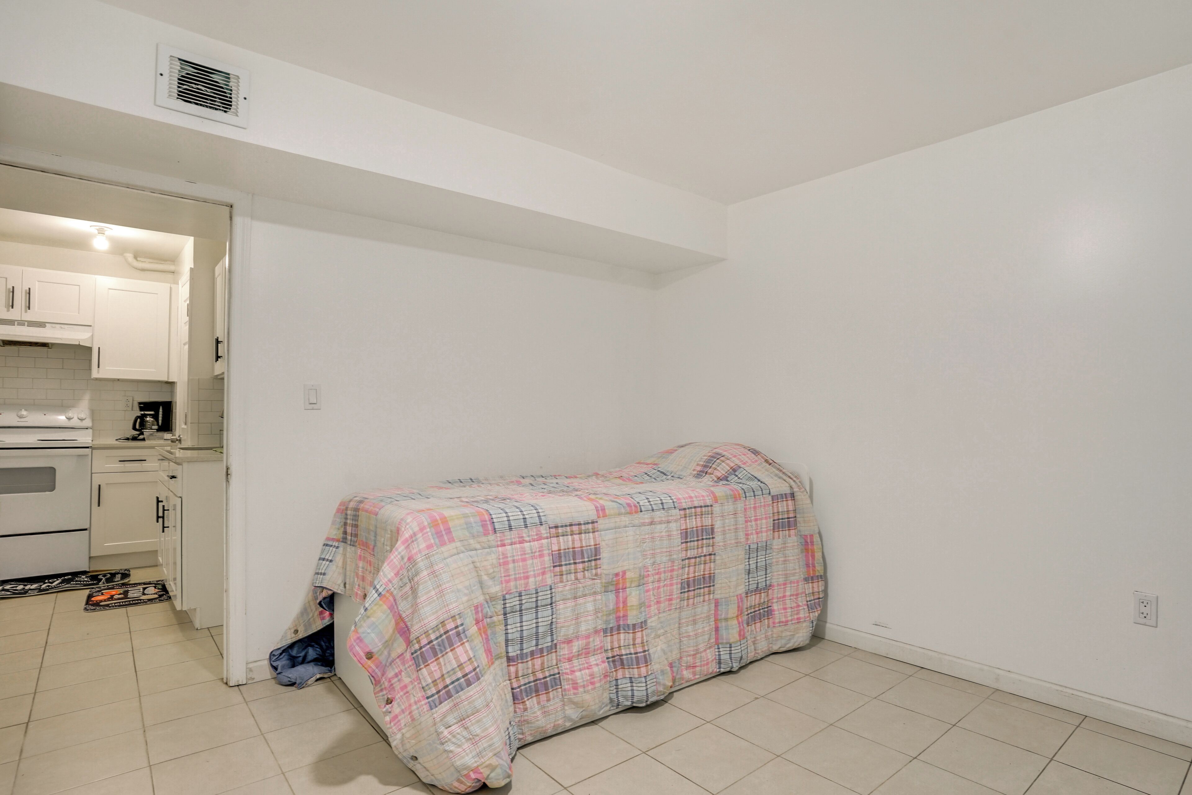 A small, clean room with a kitchenette, a bed with a plaid cover, and a door leading to another room.