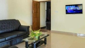 Una sala de estar con un sofá negro, una mesa de centro de vidrio, un televisor montado en la pared y una puerta de madera.