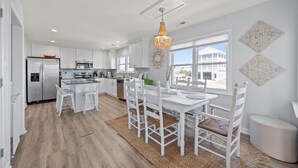 A modern kitchen with white cabinetry, a dining area with white chairs, and a large window offering a view of a house. - Bay Watch w/Elevator (Pet Friendly Home) (Morehead City)