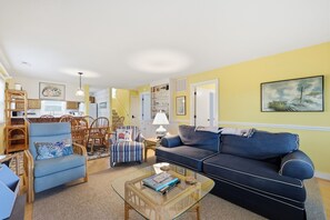 Una sala de estar con un sillón azul, un sofá azul, una mesa de centro de vidrio y una zona de comedor con sillas de madera y una mesa.