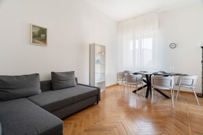 Ein modernes Wohnzimmer mit einem grauen Sofa, einem Esstisch mit Stühlen, einem Bücherregal und einem Gemälde an der Wand.