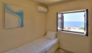 A single bed with a white mattress and pillow, a wall-mounted air conditioner, and a window with an open view of the sea and lounge chairs.