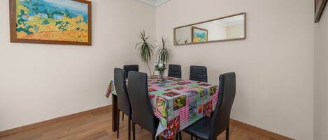 A dining area with a table covered by a quilt, surrounded by chairs, a vase with plants, and a painting on the wall.