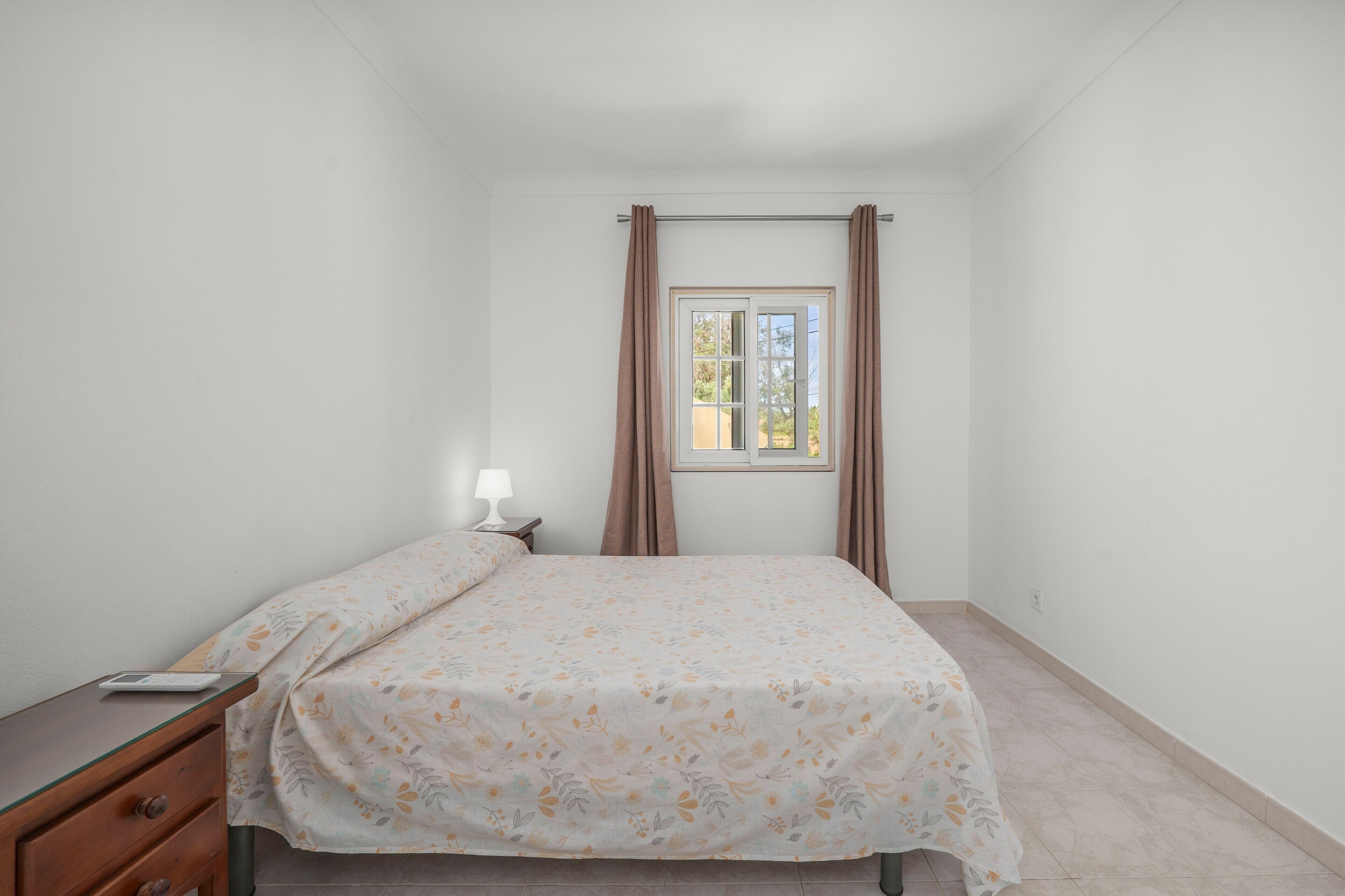 Un dormitorio con cama, mesita de noche, lámpara, ventana con cortinas y vistas al exterior.