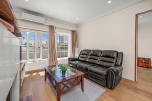 Una sala de estar con una mesa de centro de vidrio, un sofá de cuero, un aparador de madera y una ventana con cortinas.