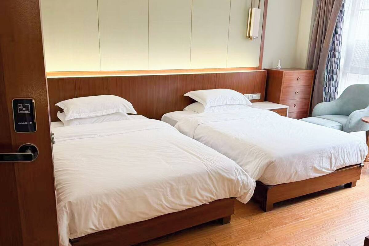 A hotel room with two beds, wooden flooring, a wooden headboard, a small desk, a chair, and a mirror.