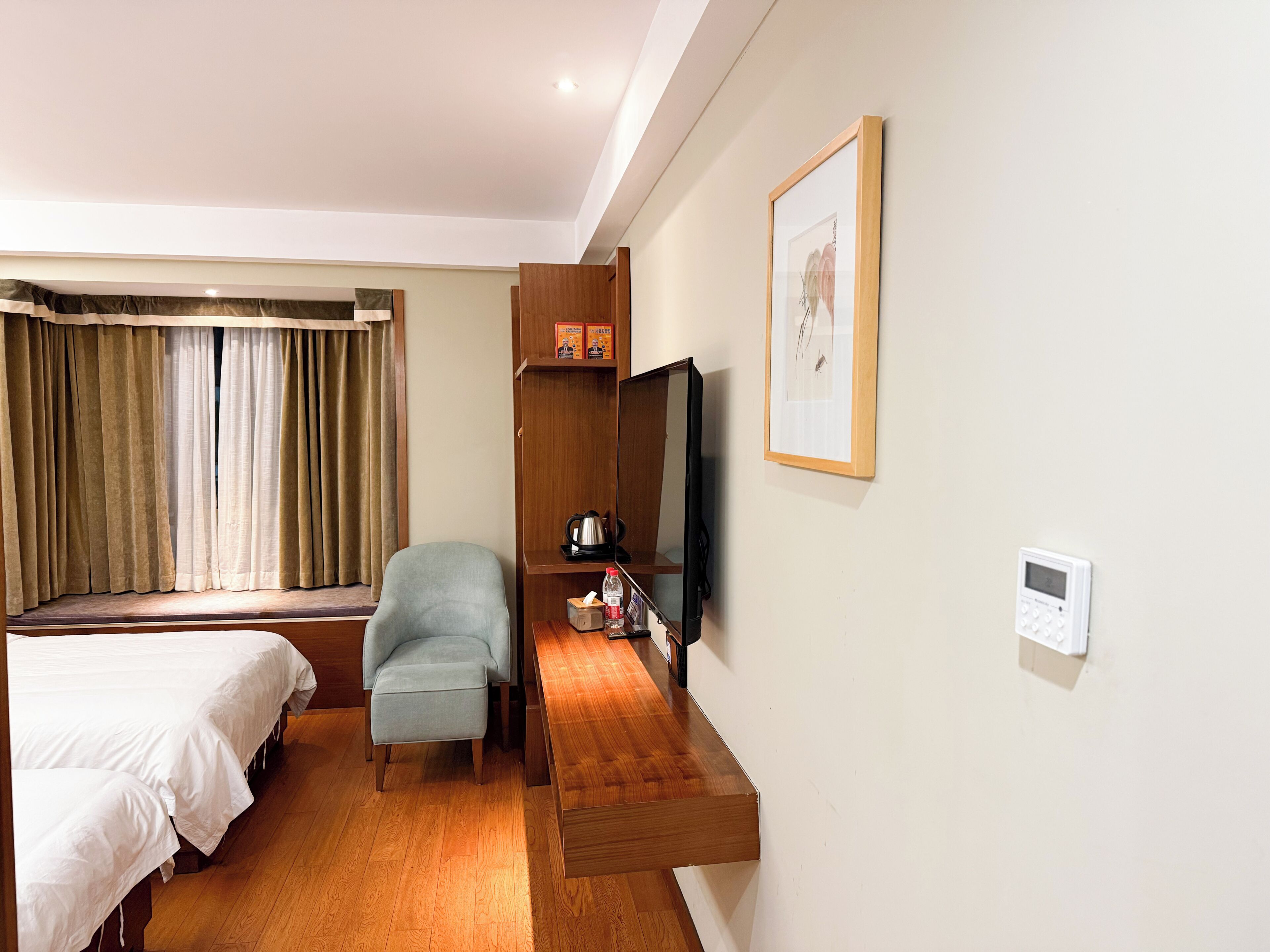 Quarto de hotel com cama, cadeira, televisão, prateleira com objetos e termostato fixado na parede.