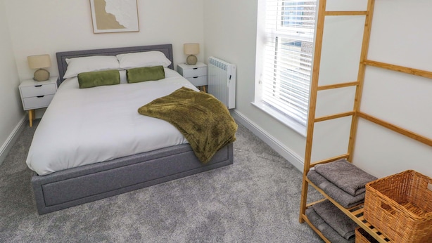 Un dormitorio ordenado con cama, mesitas de noche, un radiador, una ventana con persianas y un estante de madera con toallas dobladas.
