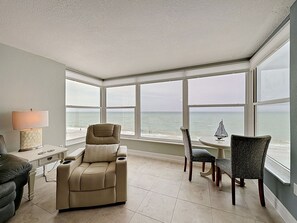 Ein Zimmer mit einem großen Erkerfenster, einem beigen Sofa, einem Esstisch mit Stühlen und einer Lampe.