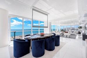 Ein modernes Wohnzimmer mit großem Meerblick, einem Essbereich mit rundem Tisch und einer Sitzecke mit blauen Hocker.