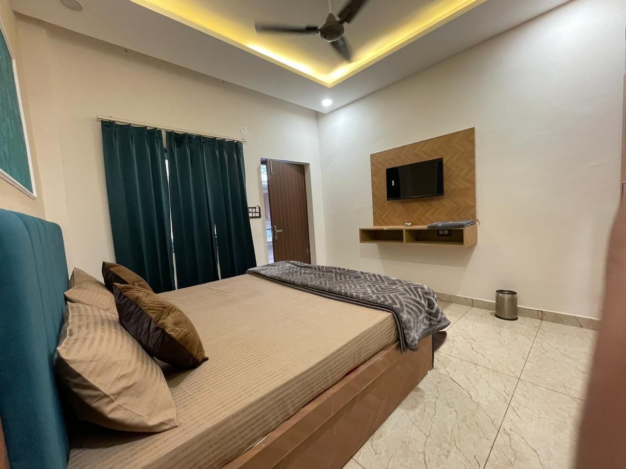 A bedroom with a bed, curtains, a ceiling fan, a mounted TV, and a small trash bin.