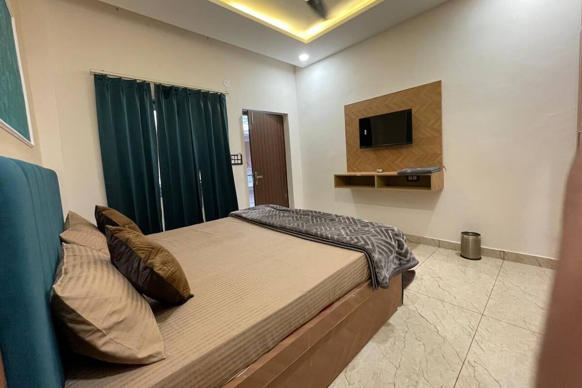 A bedroom with a bed, curtains, a ceiling fan, a mounted TV, and a small trash bin.
