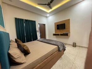 A bedroom with a bed, curtains, a ceiling fan, a mounted TV, and a small trash bin.