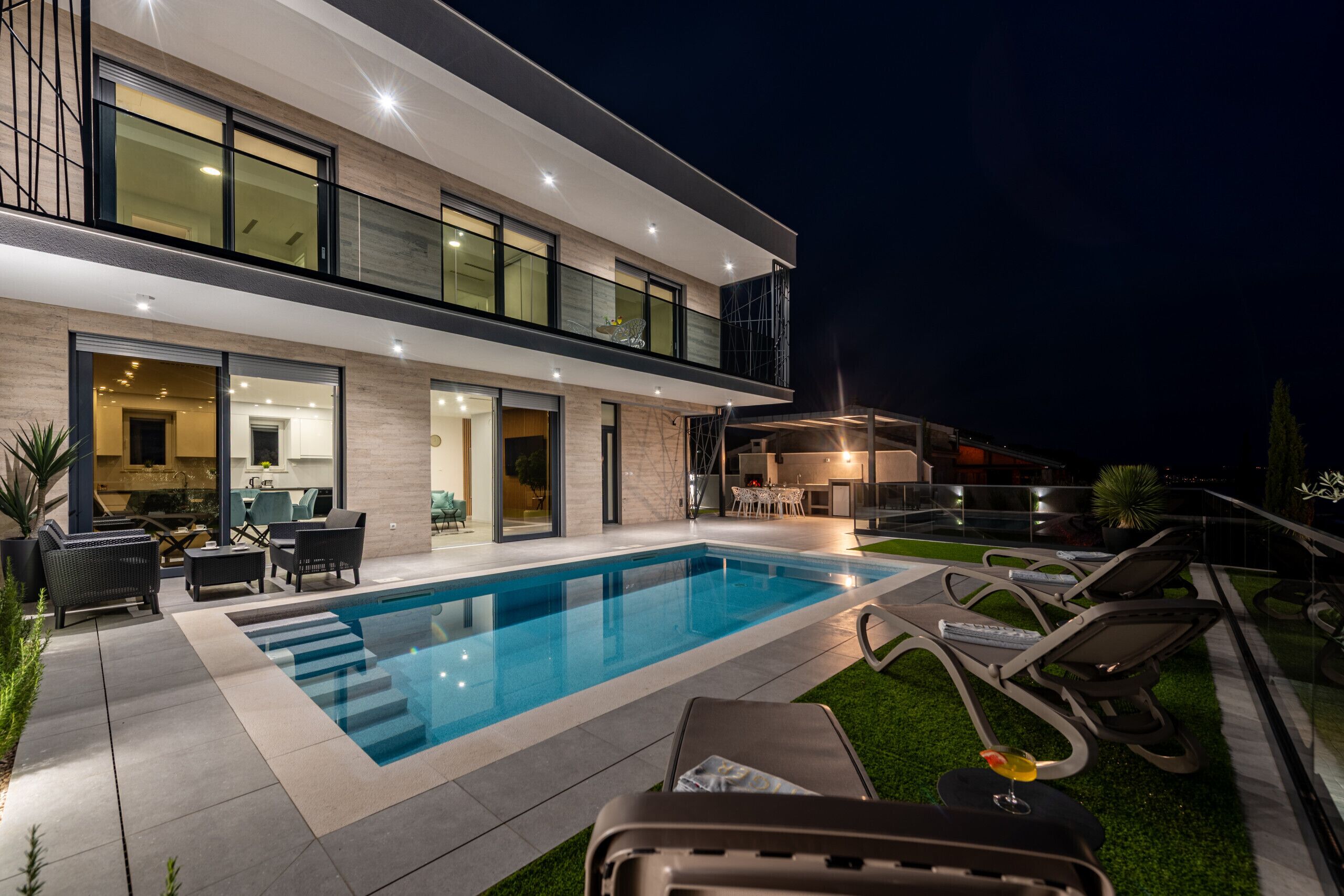 A modern two-story house with a swimming pool, outdoor seating, and a well-lit patio area.