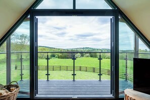 A room with a large glass window offering a view of a green field and a wooden deck. - Longdale Barn (Newtown Linford)