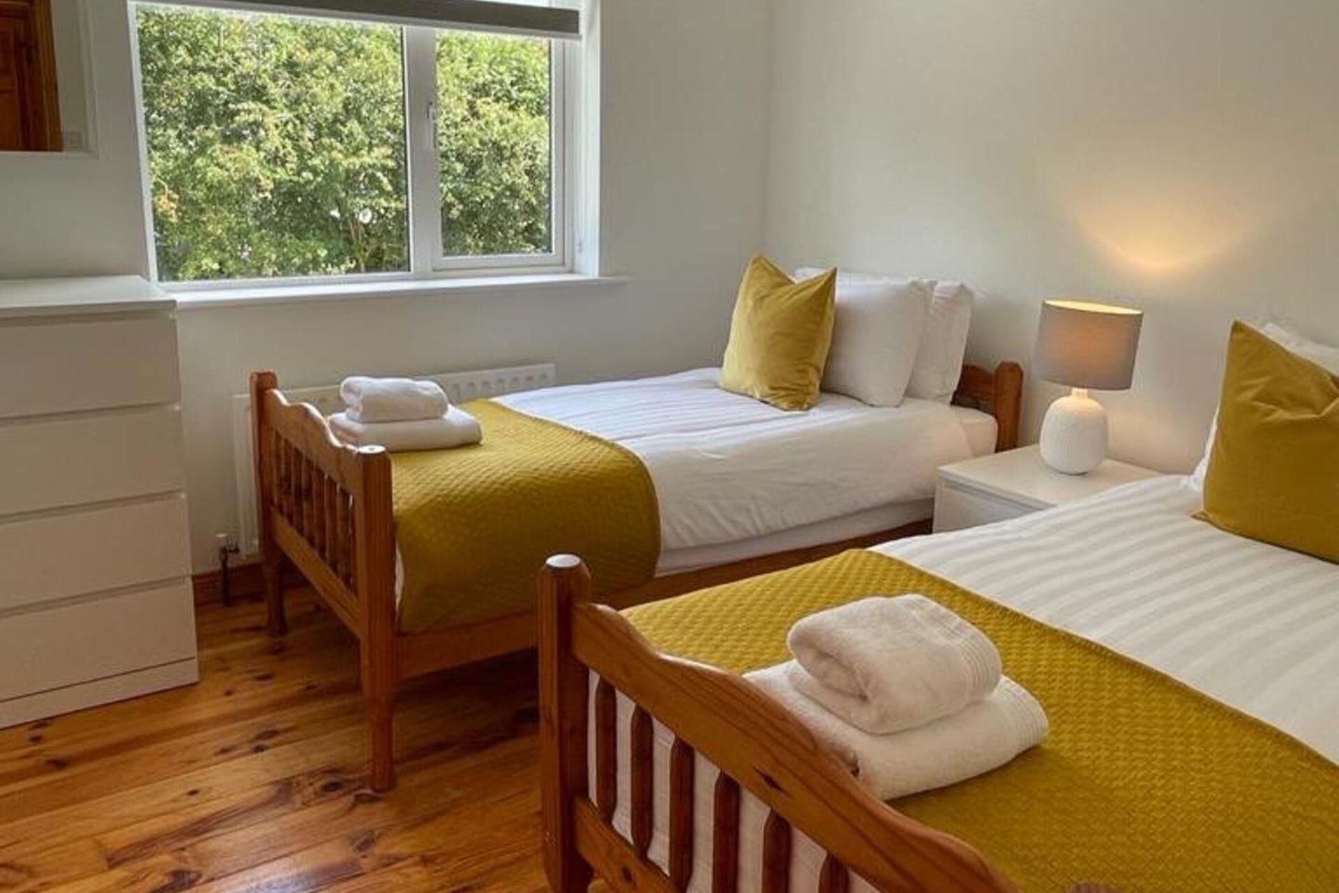 Habitación con dos camas, ventana con vista a árboles, lámpara de noche y cabecera de madera.