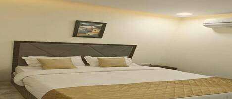 A hotel room with a bed, a nightstand, a wall-mounted air conditioner, and a framed picture on the wall.