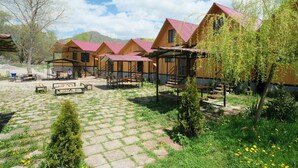 Una hilera de cabañas de madera con techos rojos, un patio pavimentado con bancos y un paisaje natural con árboles y montañas al fondo.