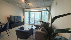 A modern living room with a flat-screen TV, sofa, chairs, and a large window with a city view. - 2BR Free Parking | Walk Wynwood & Design Dist (Miami)