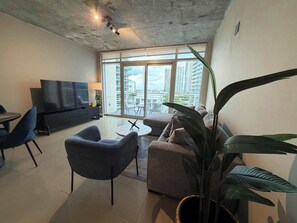 A modern living room with a flat-screen TV, sofa, chairs, and a large window with a city view. - 2BR · Free Parking · Walk Wynwood · Near Fan Fest (Miami)