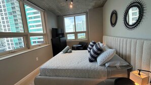 A bedroom with a large bed, a TV, a desk, and a chair. There are windows with a city view. - 2BR Free Parking | Walk Wynwood & Design Dist (Miami)