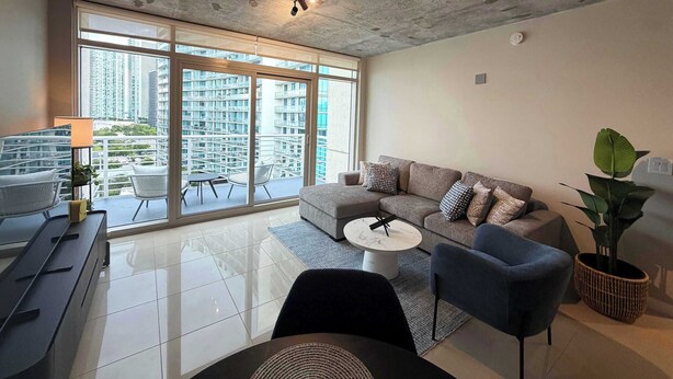 A modern living room with a sofa, chairs, a coffee table, and a balcony with a view of high-rise buildings. - 2BR Free Parking | Walk Wynwood & Design Dist (Miami)