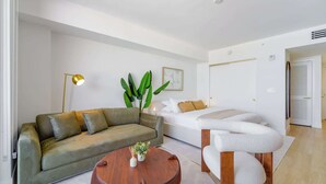 A modern living room with a sofa, a round wooden coffee table, a white chair, a floor lamp, and a large plant. - Studio in Midtown | Standard Miami - Pool & Gym (Miami)