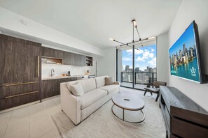 A modern living room with a sofa, coffee table, and a cityscape view through large windows. - Bright 2BR | Fitness Center | Near Bayside (Downtown)