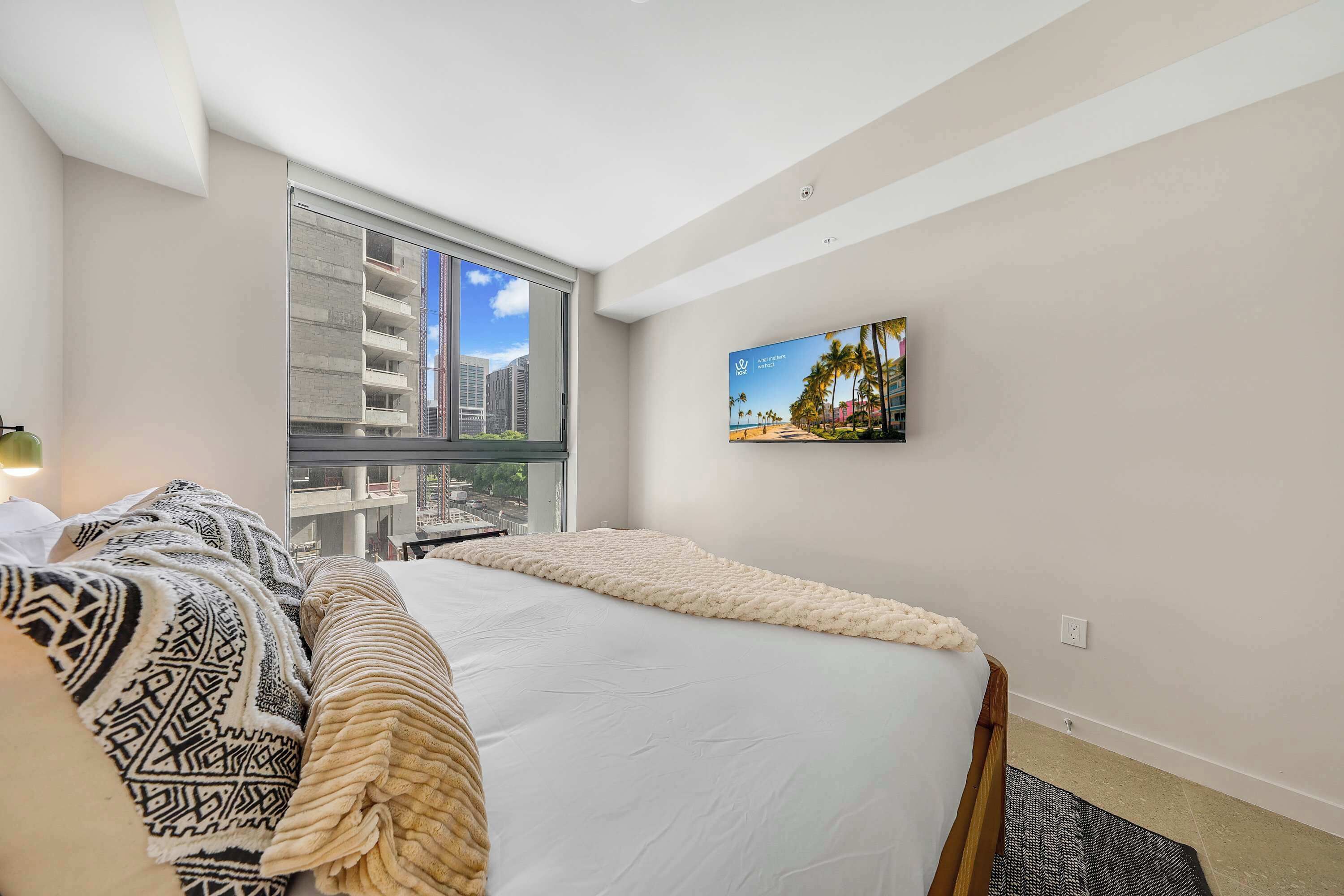 A hotel room with a large bed, a view of a cityscape through a window, and a wall-mounted TV.