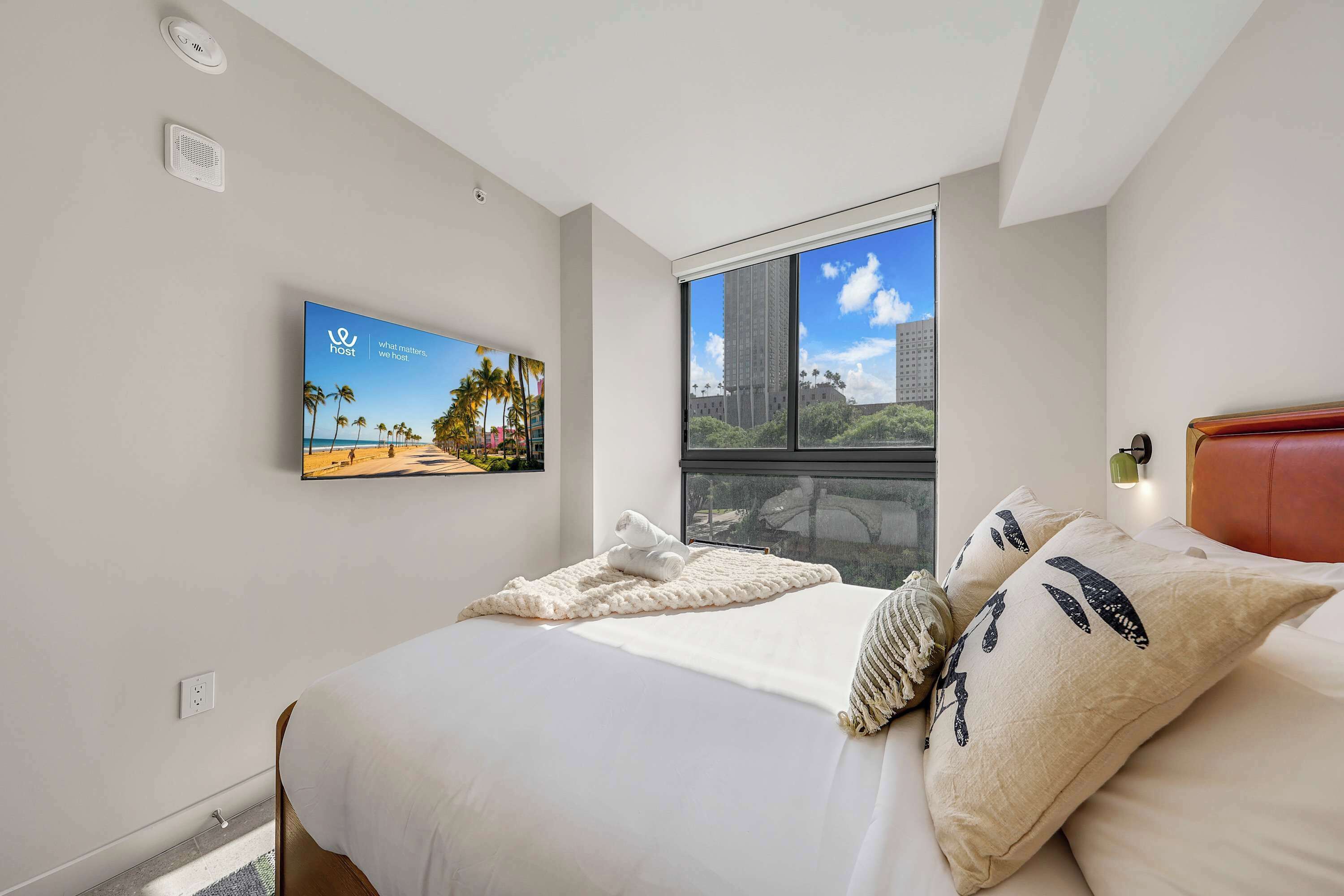 Quarto de hotel com cama, televisão fixada na parede, janela com vista para um prédio alto e termostato na parede.